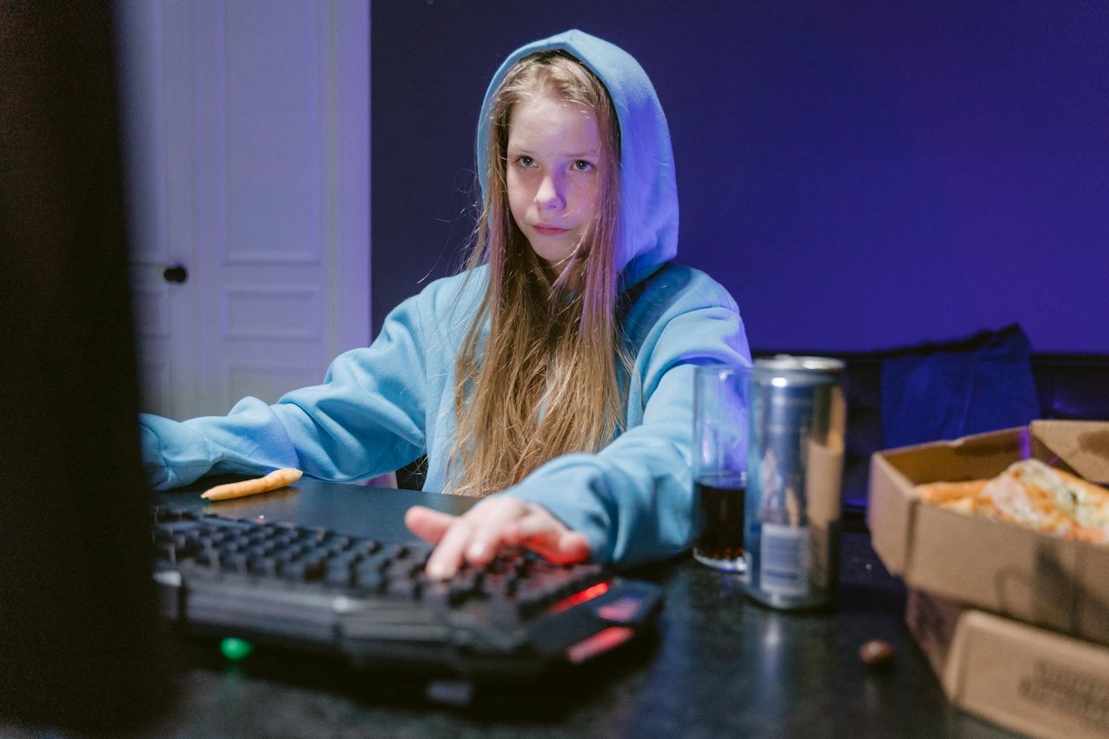Teen girl in a blue hoodie playing video games inside, surrounded by snacks and drinks.